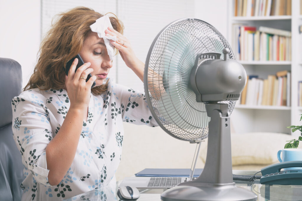 Woman suffers from hot flash while working in the office and tries to cool off by the fan
