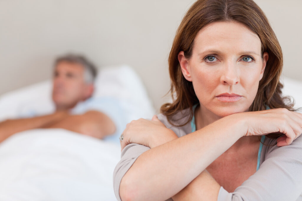 Sad woman, with low libido post hysterectomy, sitting on the bed with her husband in the background.